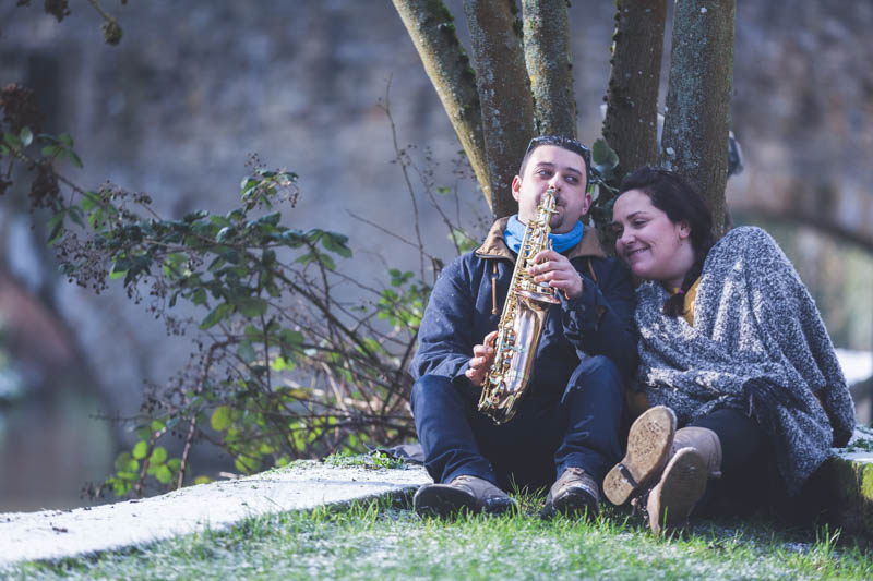 man playing saxophone under a tree next to his fiancee